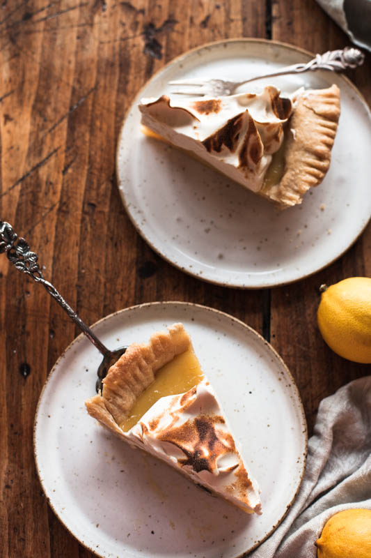 Zwei Kuchenstücke Lemon Meringue Pie / Zitronenkuchen auf zwei weißen Tellern, die auf einer rustikalen Tischplatte stehen. 
