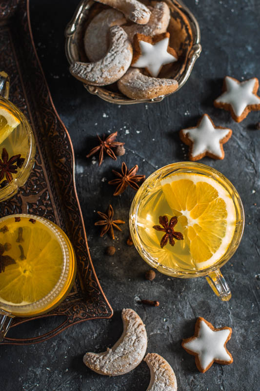 Homemade german mulled wine served in pretty crystal glasses and decorated with orange slices and star anise. Christmas cookies are spread around the glasses and served in a small bowl.