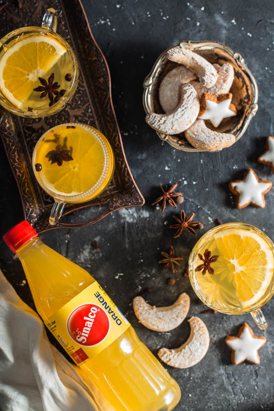 A festive setup on a dark gray surface containing three crystal glasses filled with mulled white wine, a bowl of Christmas cookies and a bottle of orange lemonade from Sinalco